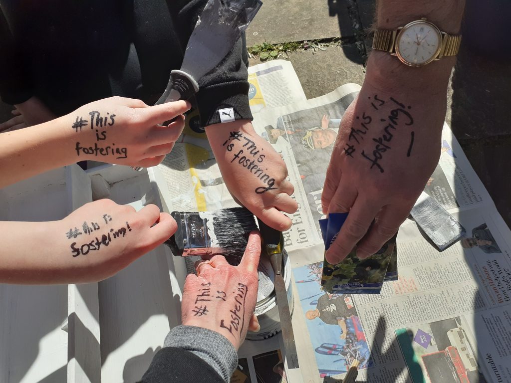 A family of 5 show their hands with #ThisIsFostering written on them. They are all holding a tool or gardening implement to show the family all working together.